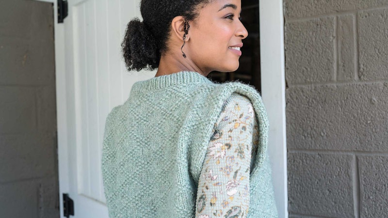 A woman wearing a textured slipover looks into the distance. She is smiling.