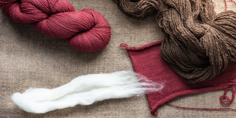 Clockwise, from top left: a red skein of yarn from Sincere Sheep, a brown skein of handspun yarn, a handknitted swatch, and undyed white Cormo wool. All rest on a handwoven fabric with Cormo weft.