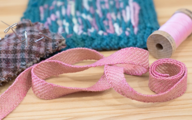 A pink handwoven band with pink thread, a plaid pincushion, and a turquoise and pink swatch on a wooden background.