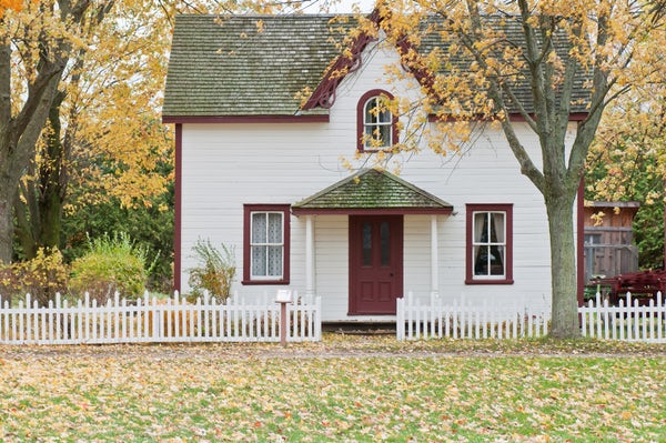 White two-story home with red door and trim surrounded by trees with yellow leaves.