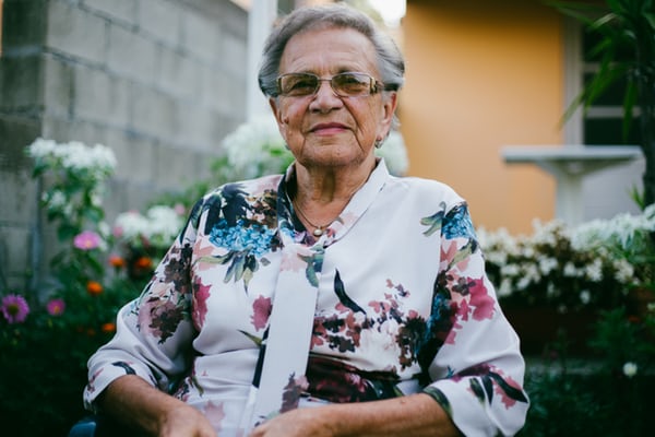 An elderly woman seated in front of a garden with a yellow house in the background