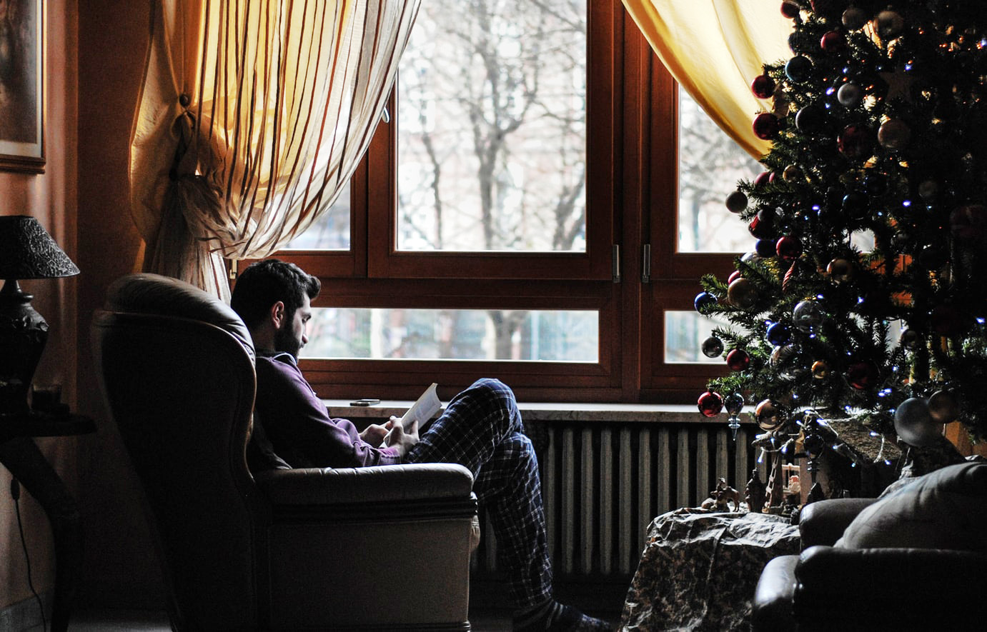 man reading in a chair by a window and Christmas tree