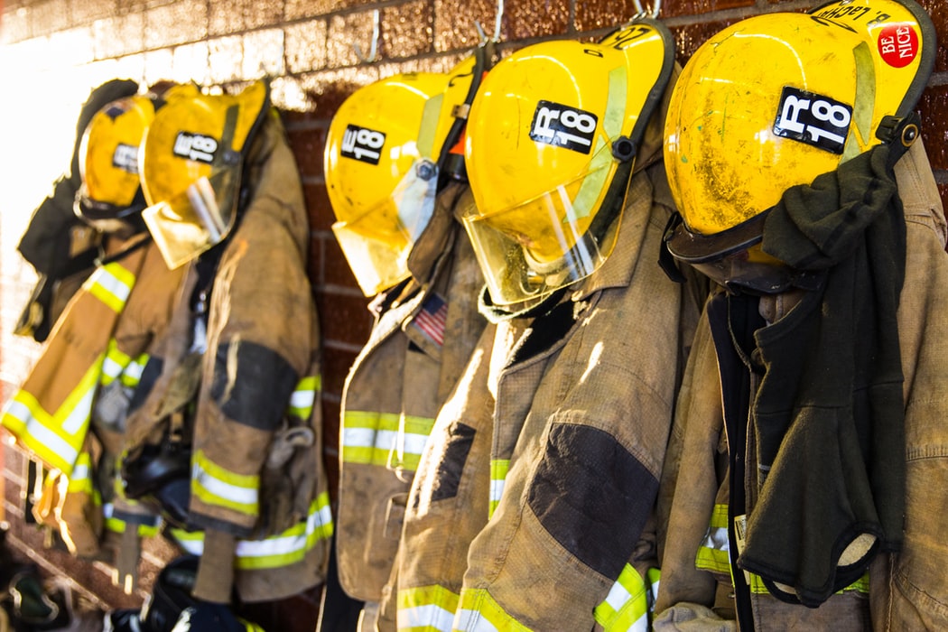 Fire department uniforms and helmets hung up on a wall in a row.