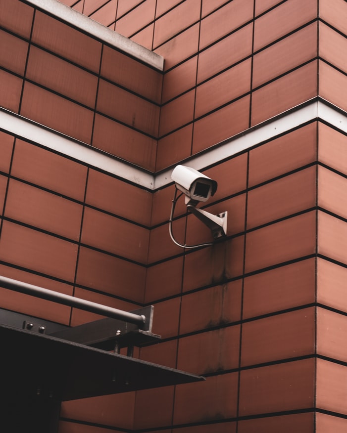A  white security camera mounted at the corner of two red orange-brown brick walls.