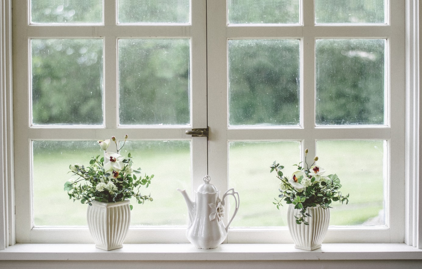 Window made up of small square glass panes. A teapot in front of it and in the middle. Two potted flower plants on each side.