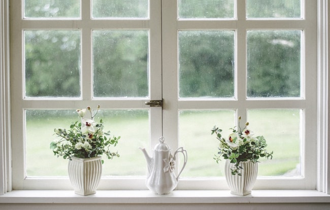 Window made up of small square glass panes. A teapot in front of it and in the middle. Two potted flower plants on each side.