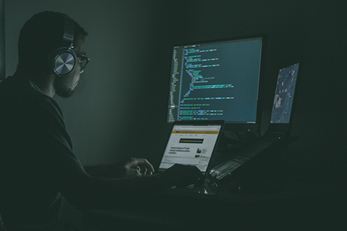 Computer programmer with headphones on in a dark room working with three screens coding a digital security program.