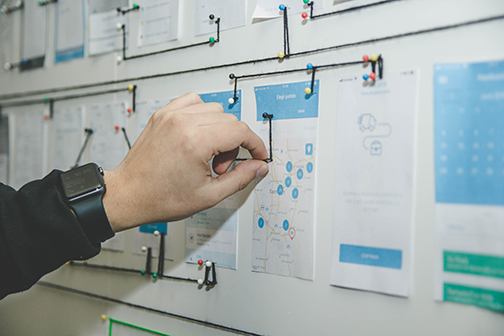 Hand connecting different pieces of the security plan with yarn and thumbtacks on a wall.