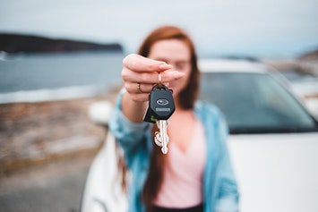 Young red haired woman holding out keys in front of her. Her white can is behind her parked near a lake. 