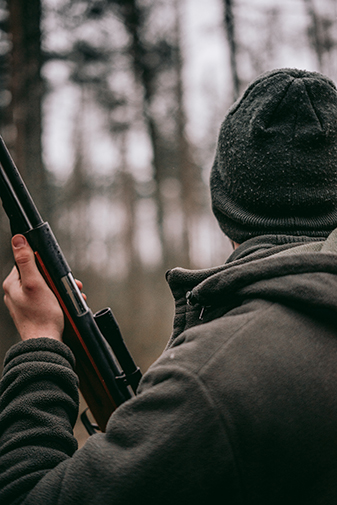 Hunter turned away from camera wearing a gray coat and beanie while wielding a hunting rifle in the forest. 