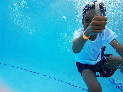 Kid in swim suit and white shirt with orang wristband giving thumbs up underwater