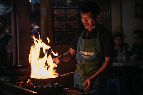 A cook is cooking food in a pan. The pan is filled with a foot high fire from burning oil.