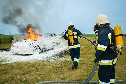 Two Firefighters Putting Out a Car Fire with a fire hose. The car is burning and the outside is covered with a white foam. 