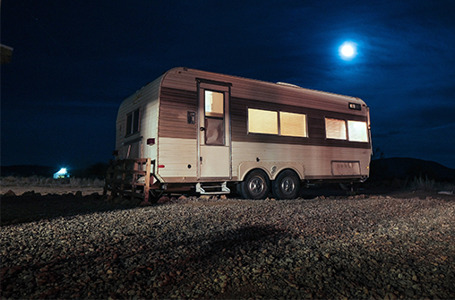 RV parked on small rocks. The lights are on inside and the moon is high in the sky.