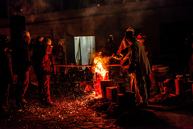 Several men lit by the light of the molten metal they are pouring. The metal is on fire.