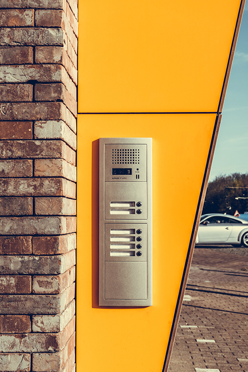 Doorbell for apartments mounted on a yellow wall and brick wall. Car in background.