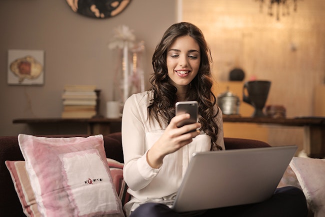 Woman sitting on a couch accessing the the app for her wireless security system. She is also on her laptop.