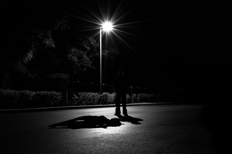 Criminal standing over victim, silhouetted from the light of a white streetlight. 