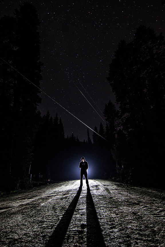 Criminal standing in dark clothing at night backlit by a spotlight.