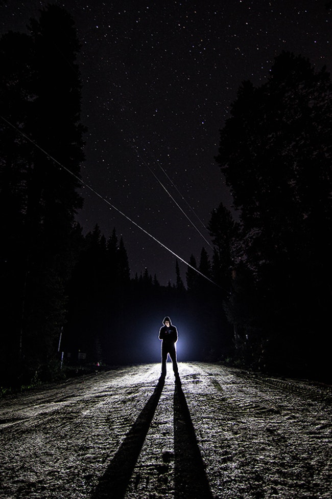 Criminal standing in dark clothing at night backlit by a spotlight.