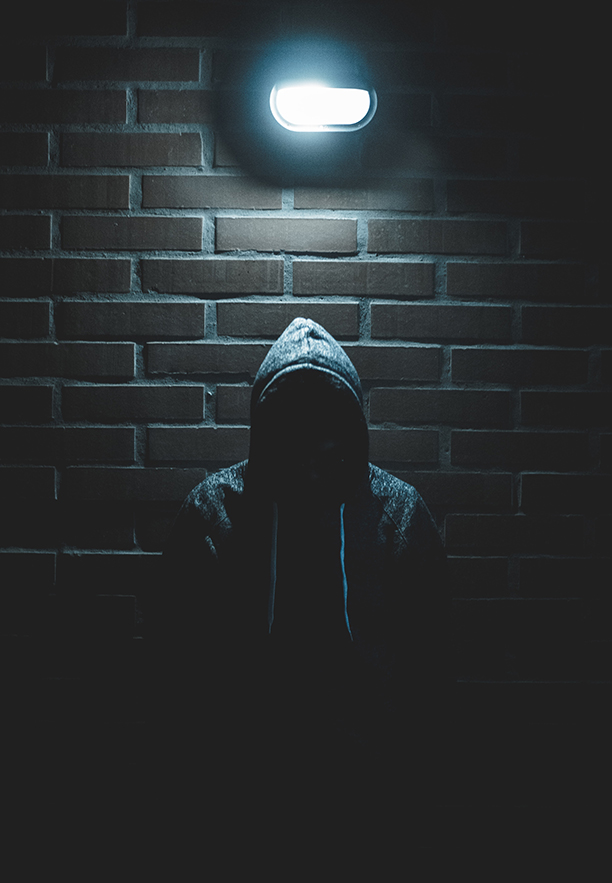 Burglar In a hoody (face obscured) at night underneath a white light sticking out of a brick wall.