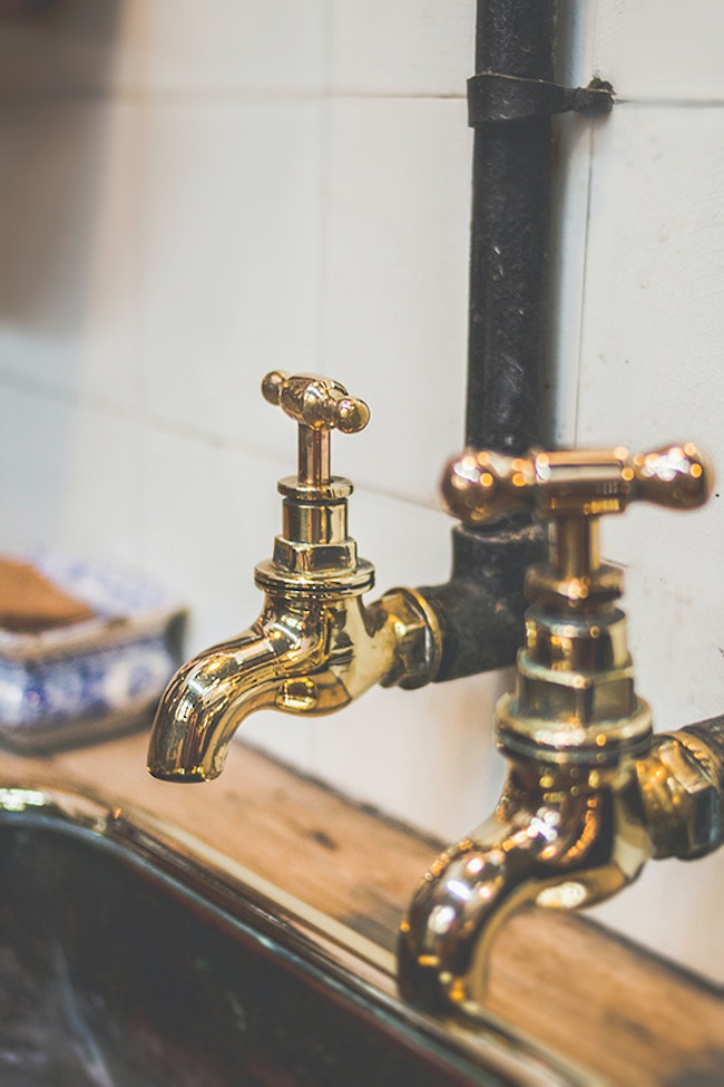 Two gold colored faucets above a sink with a connected pipe running up the tiled wall.