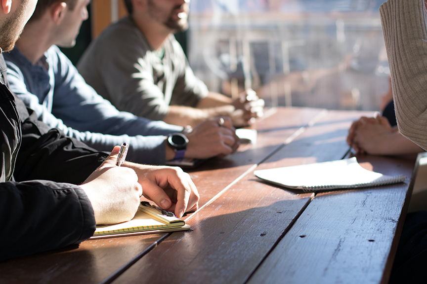 Employees At A Business Meeting taking notes as sunlight pours onto the wooden table.