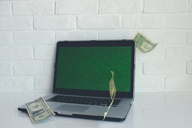 laptop with code on it and dollar bills surrounding it representing the stolen money as a result of an information breach.