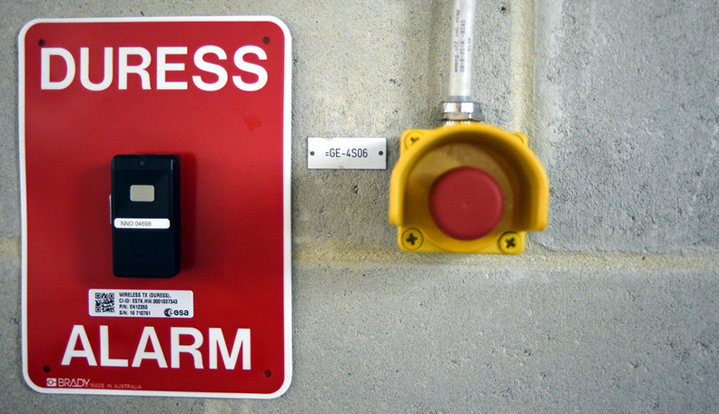 Duress alarm mounted on a cement wall accompanied by a sign saying duress alarm.