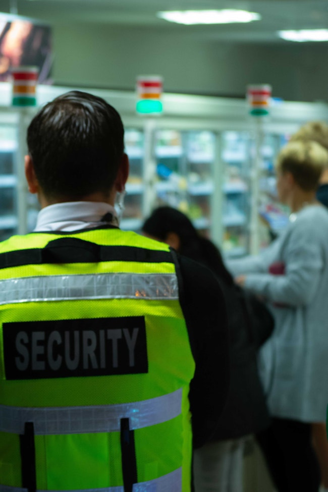 A Security Guard in a neon yellow vest looking at a room with people looking at products