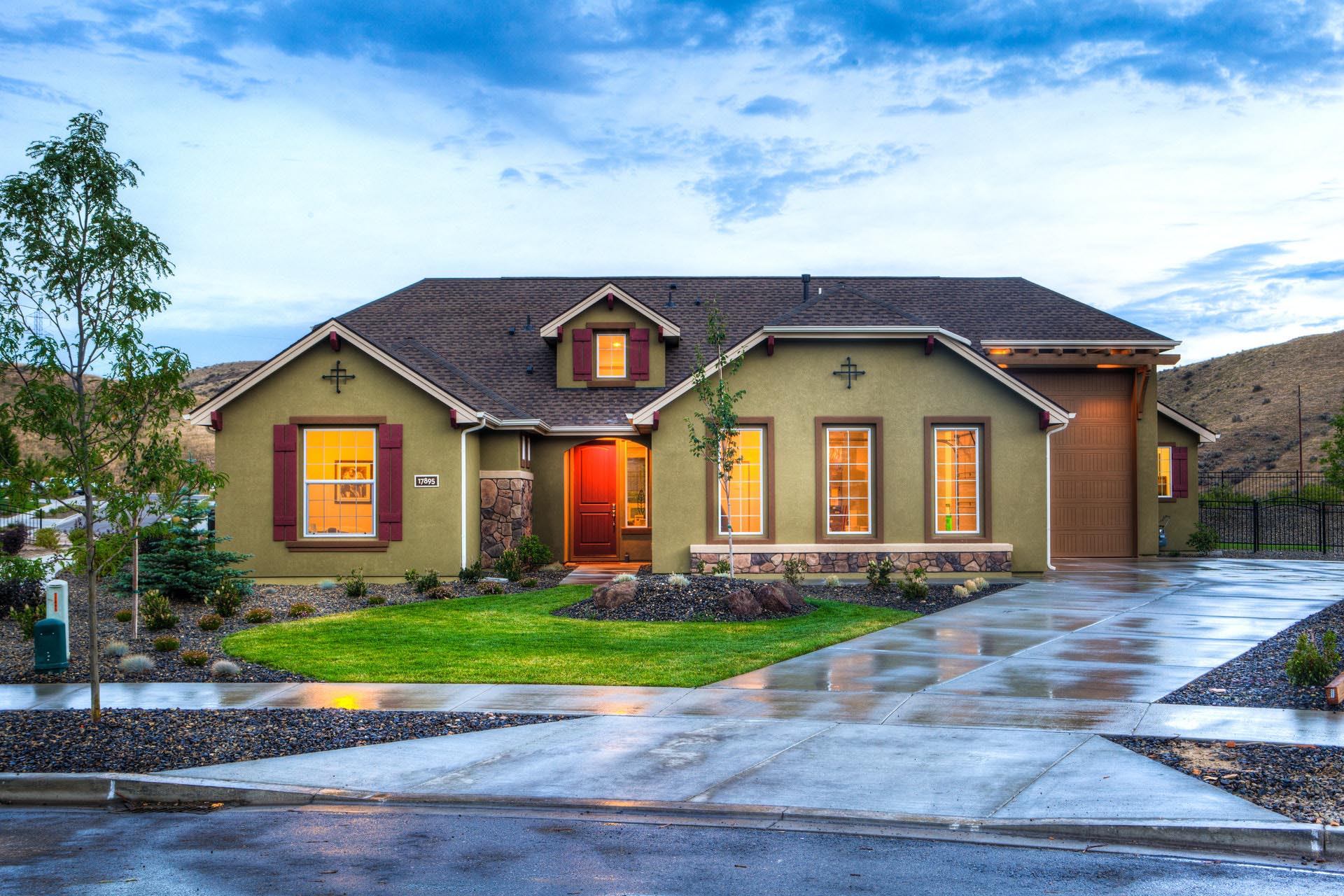 A house painted green with a red door, warm light glows from inside and reflects off of the wet concrete driveway