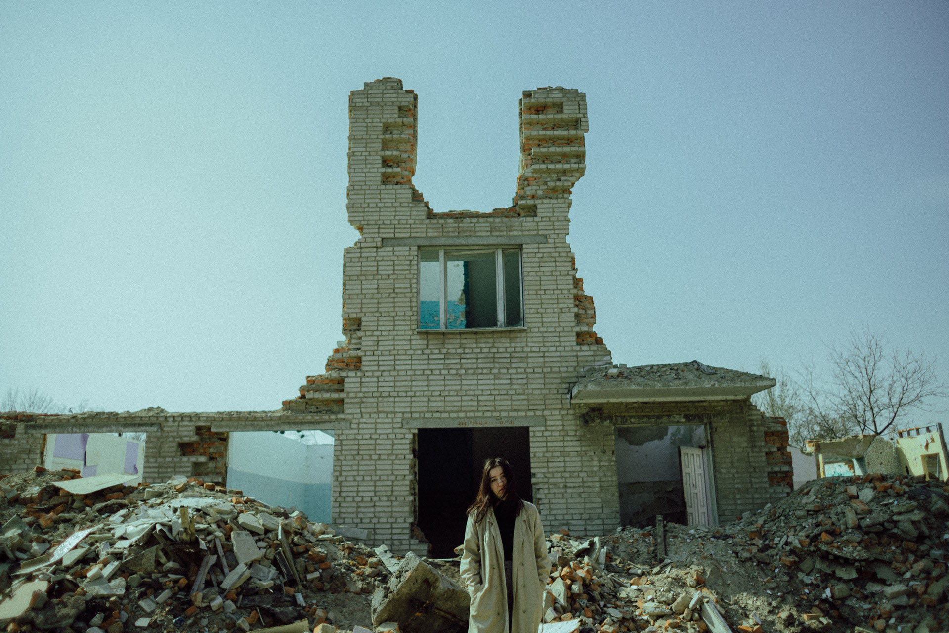 A woman in a brown cardigan is standing in front of a destroyed brick building.