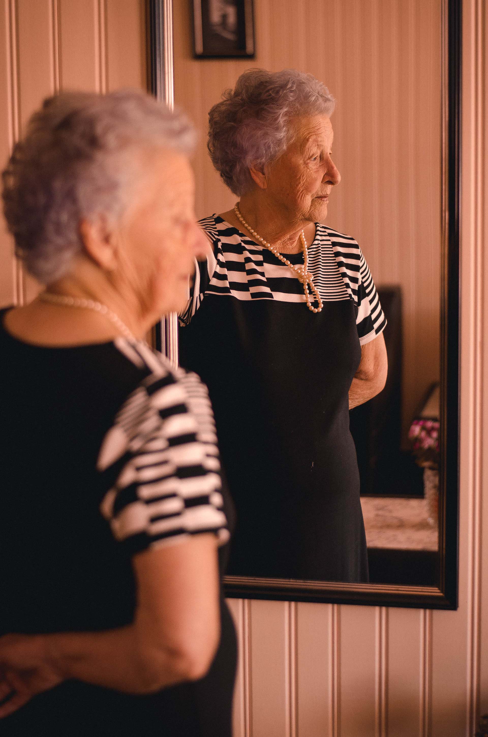 An elderly woman standing with a black dress and gold necklace in front of a mirror