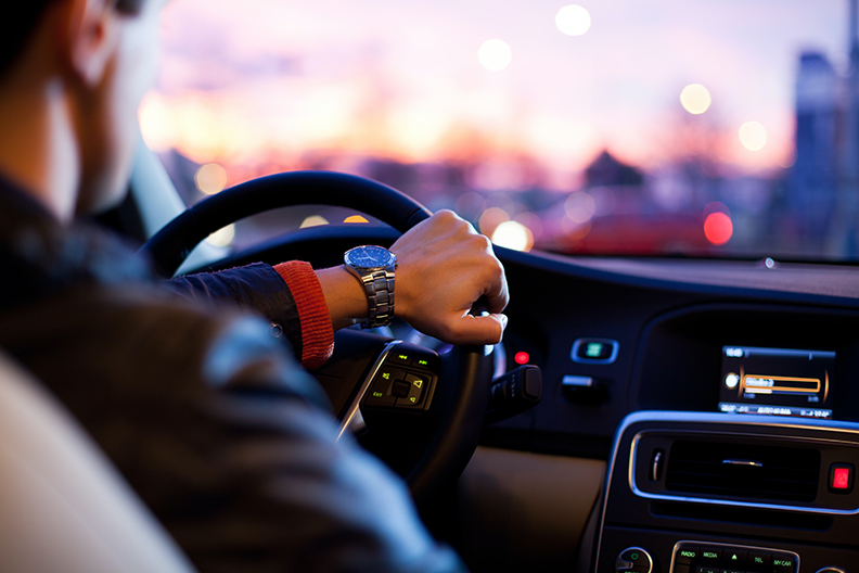 Man in a suit driving at sunset with one hand on the wheel.