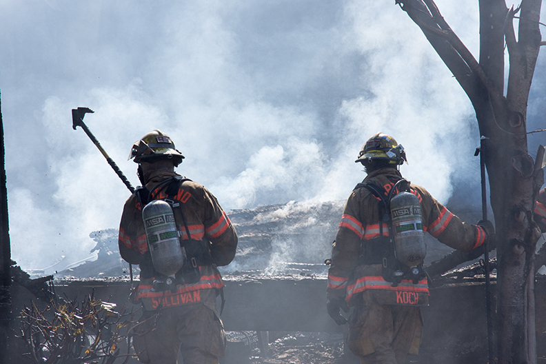 Two firefighters with oxygen tanks and axes walk through a smoking ditch.