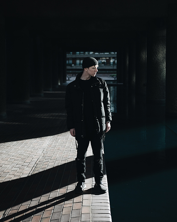 Man dressed in dark clothes, standing under a bridge