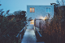 House surrounded by coastal plants. Warm light pours from a porch light and from lights inside the windows.
