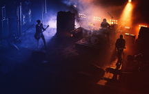 Three band members stand in the smoky darkness only lit from behind by different colored lights.
