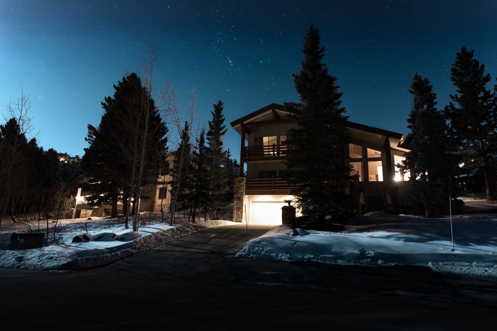 A photo of a wooden house, trees, and snow at dusk. Warm light comes from both inside and outside of the house. 
