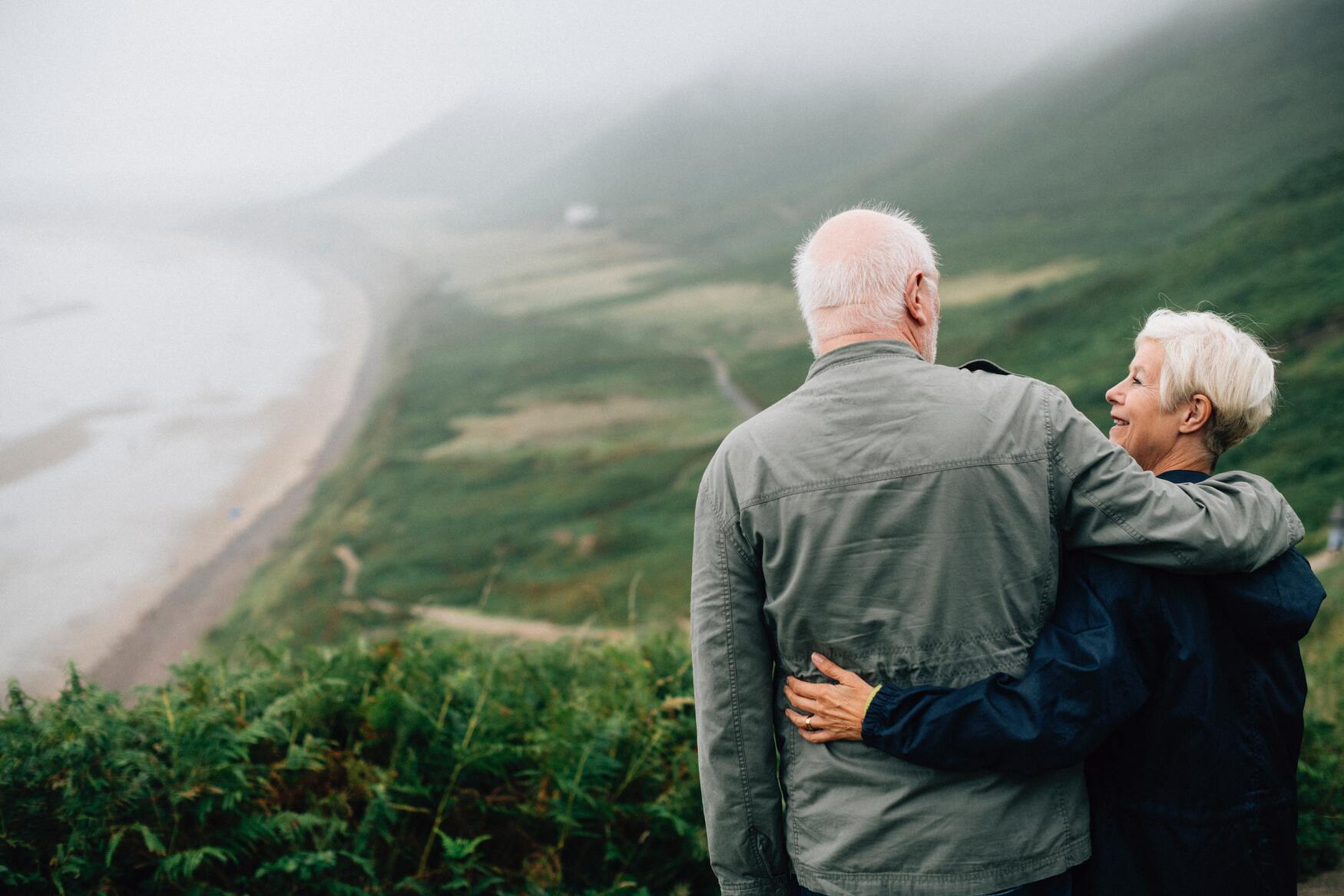 Older couple smiling at one another with one arm around the other. The background is a beach with a green shore.