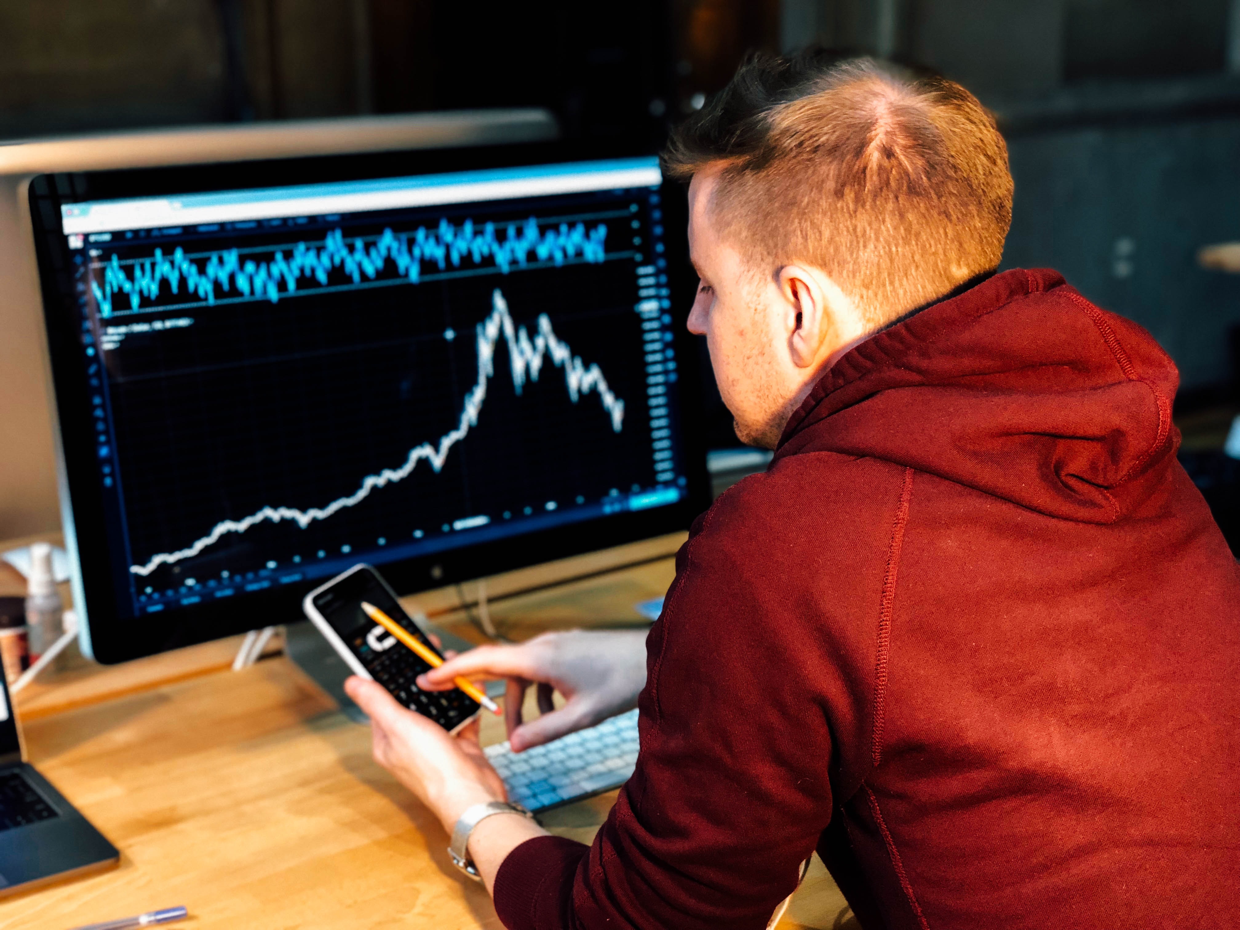 Man in red hoodie scrolling his phone in front of a computer screen with a graph chart.