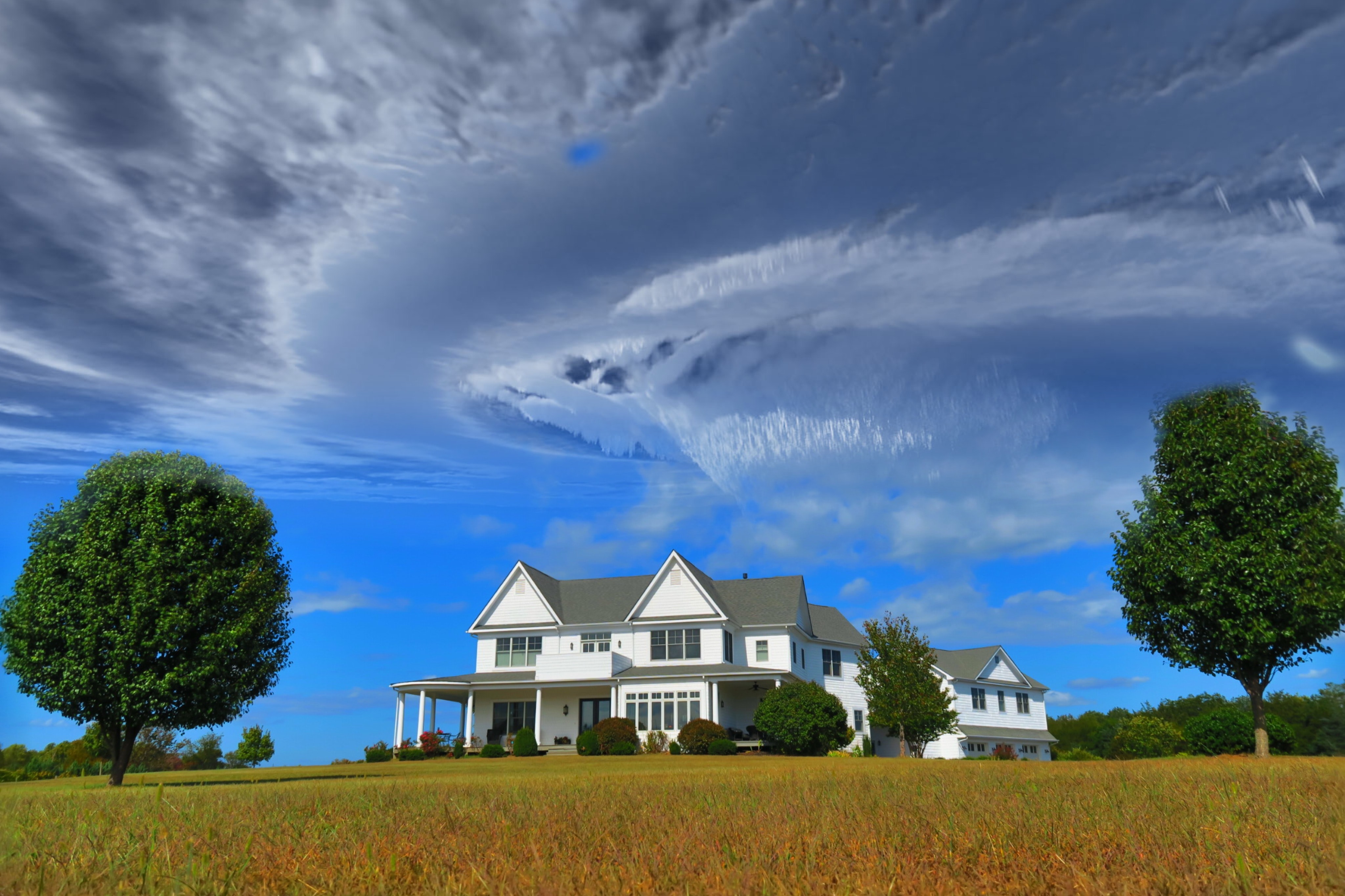 White country home on property with two trees and swirling clouds.