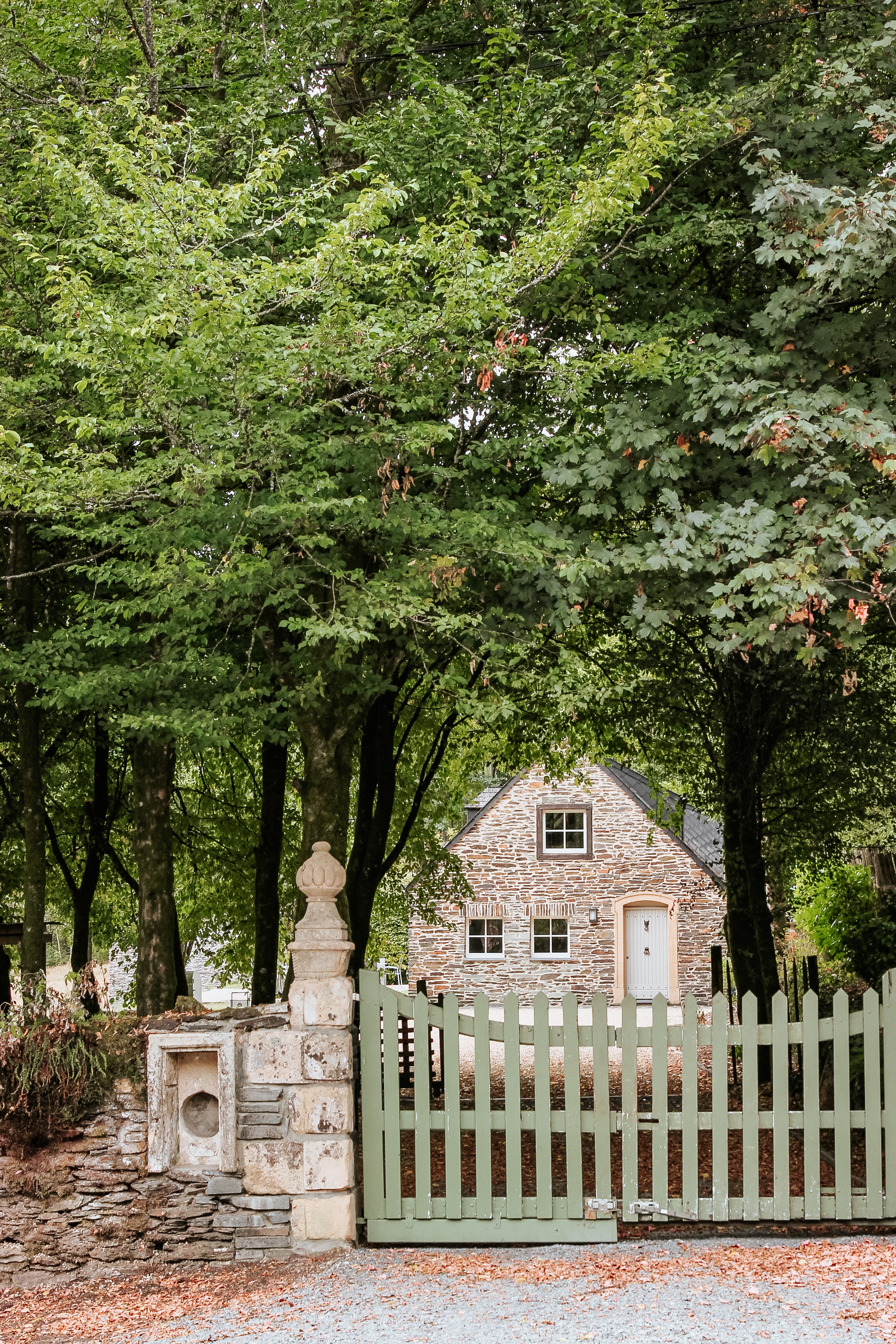 A stone house surrounded by trees with a green gate and stone fence in front.