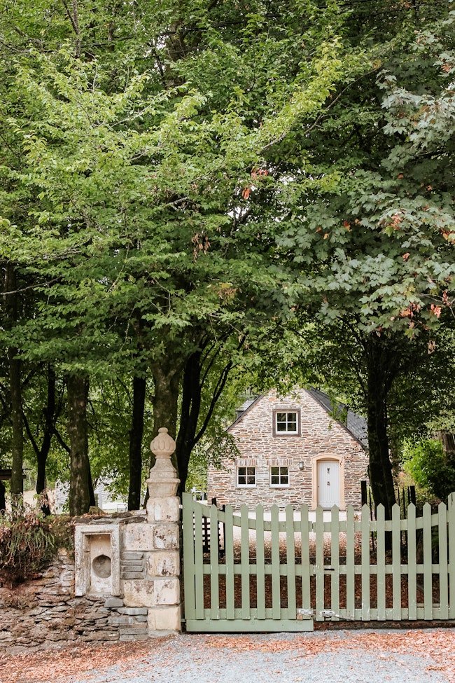 A stone house surrounded by trees with a green gate and stone fence in front.