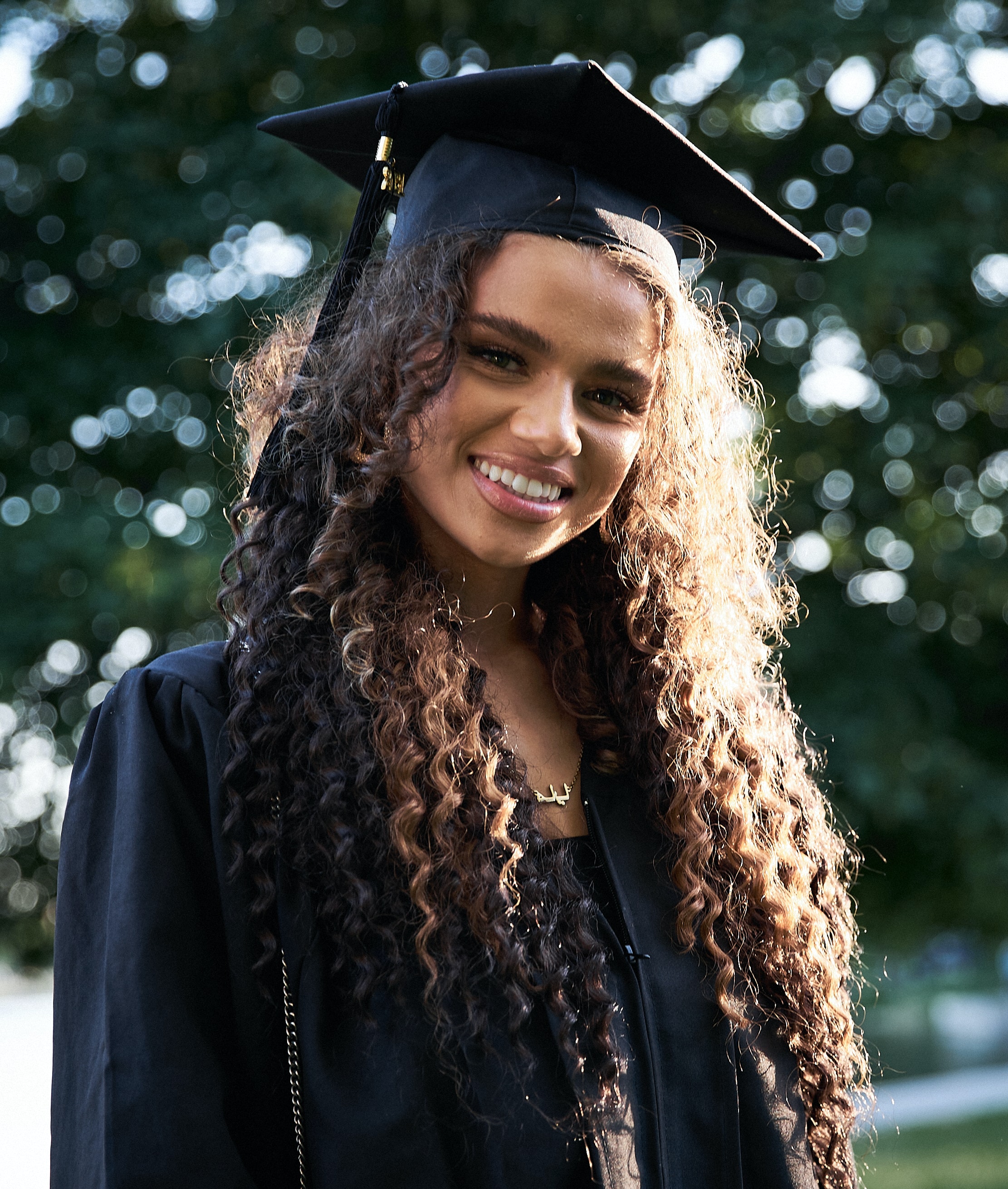 Young woman with dark curly hair dresses in a graduation cap and gown.