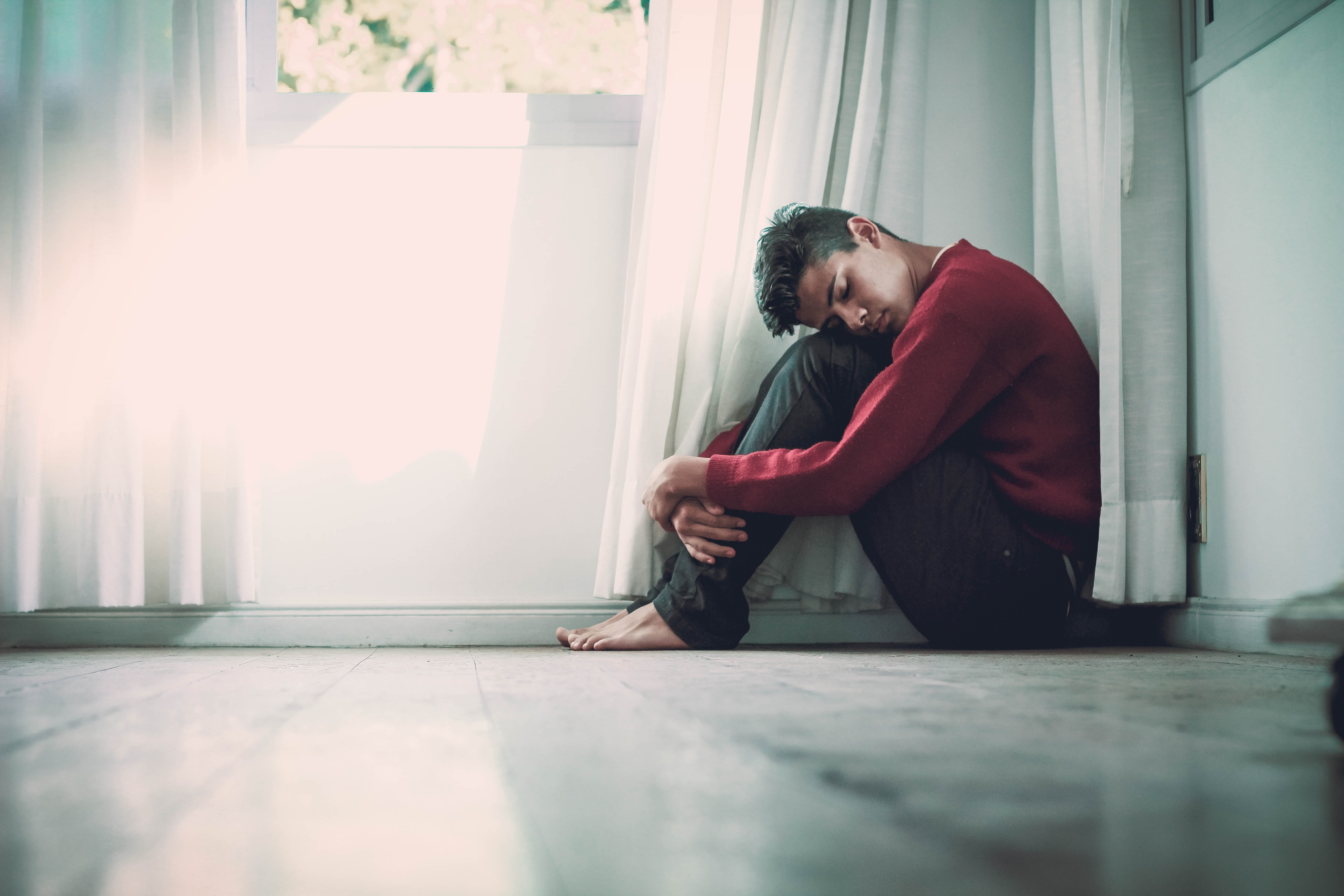 Young man on the floor with his arms wrapped around his legs, his head leaning on his knees.