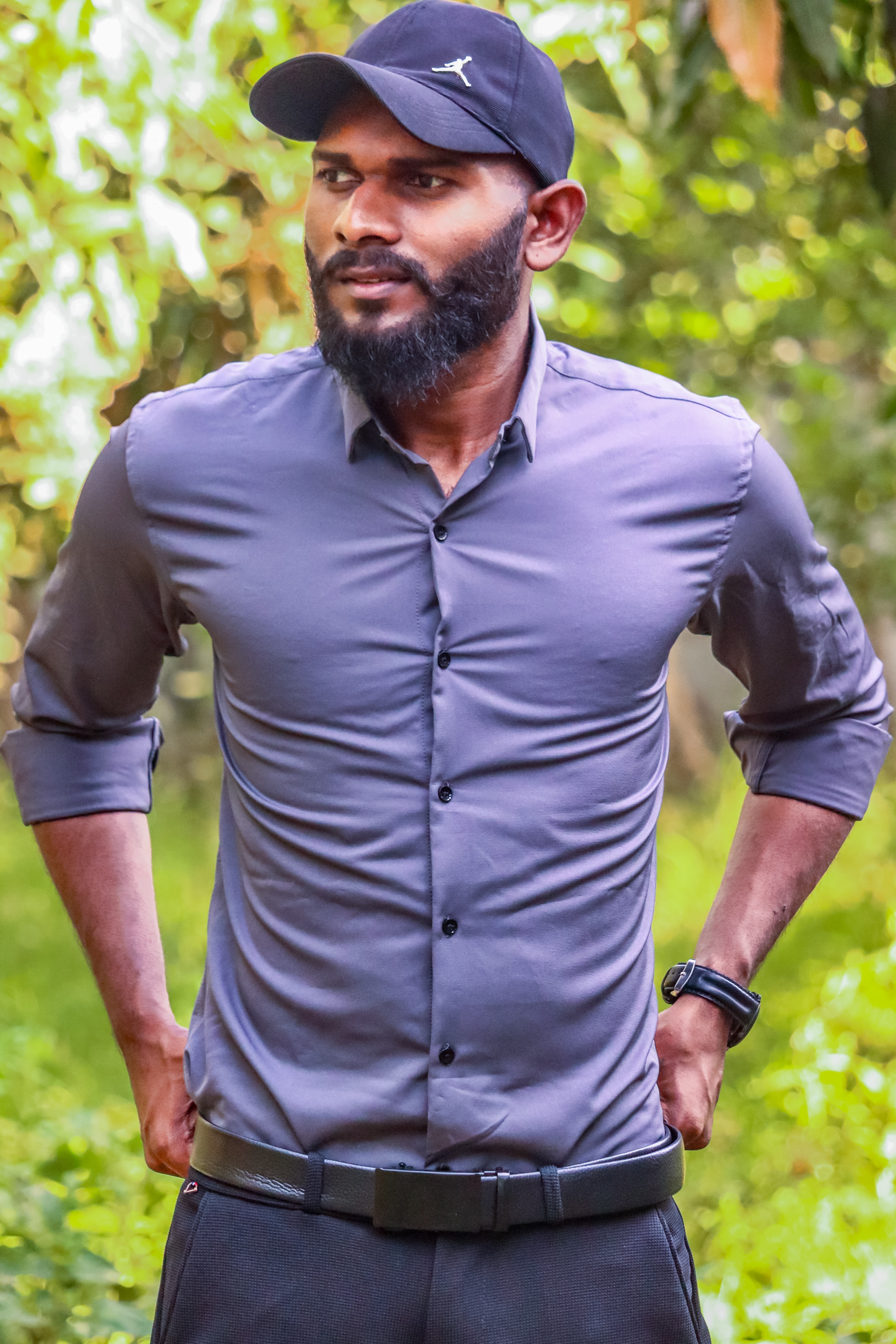 Man with a purple shirt wearing a hat and a belt outside with greenery in the background.