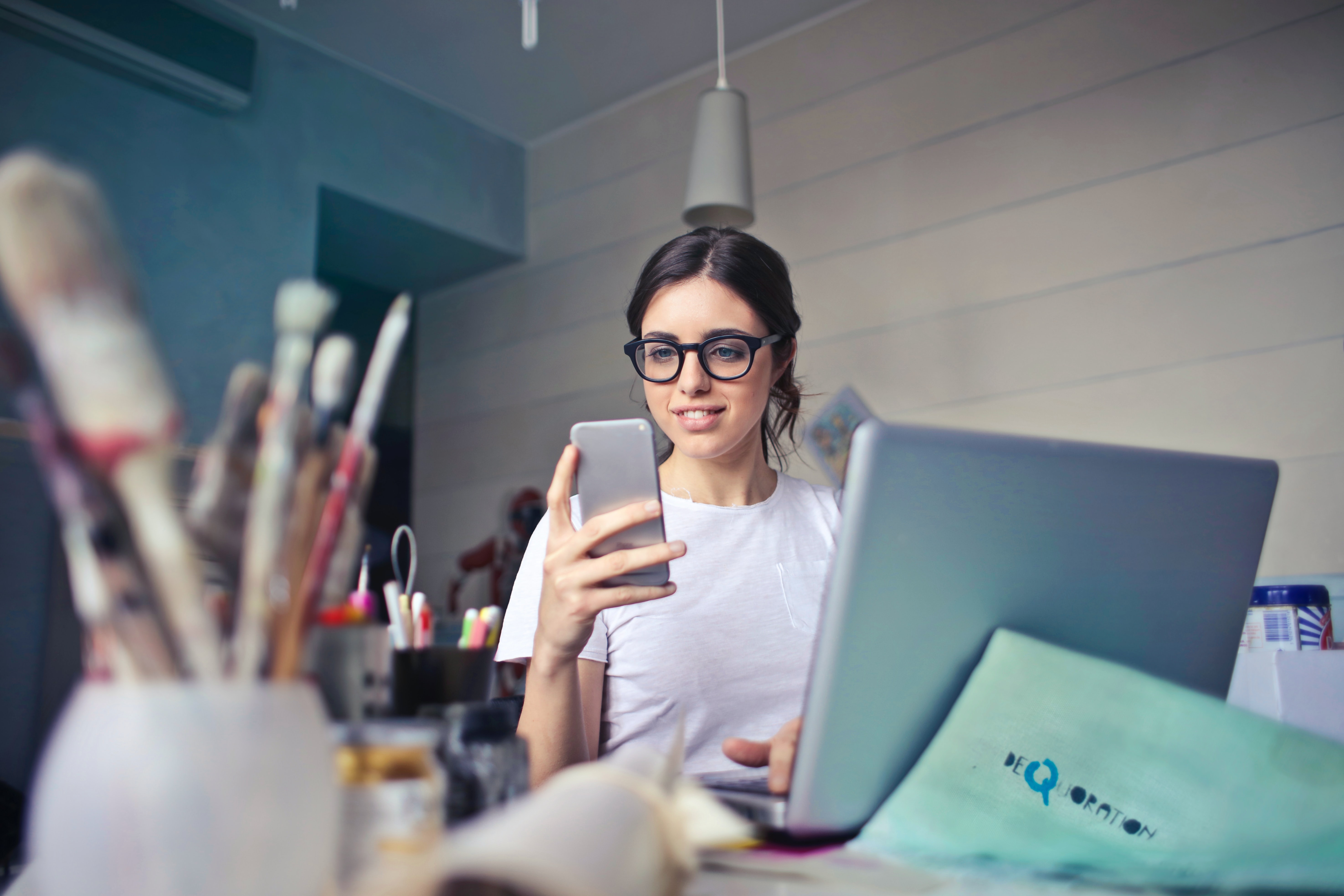 Woman wearing glasses looking on her phone surrounded by laptop and paint brushes in her home studio.