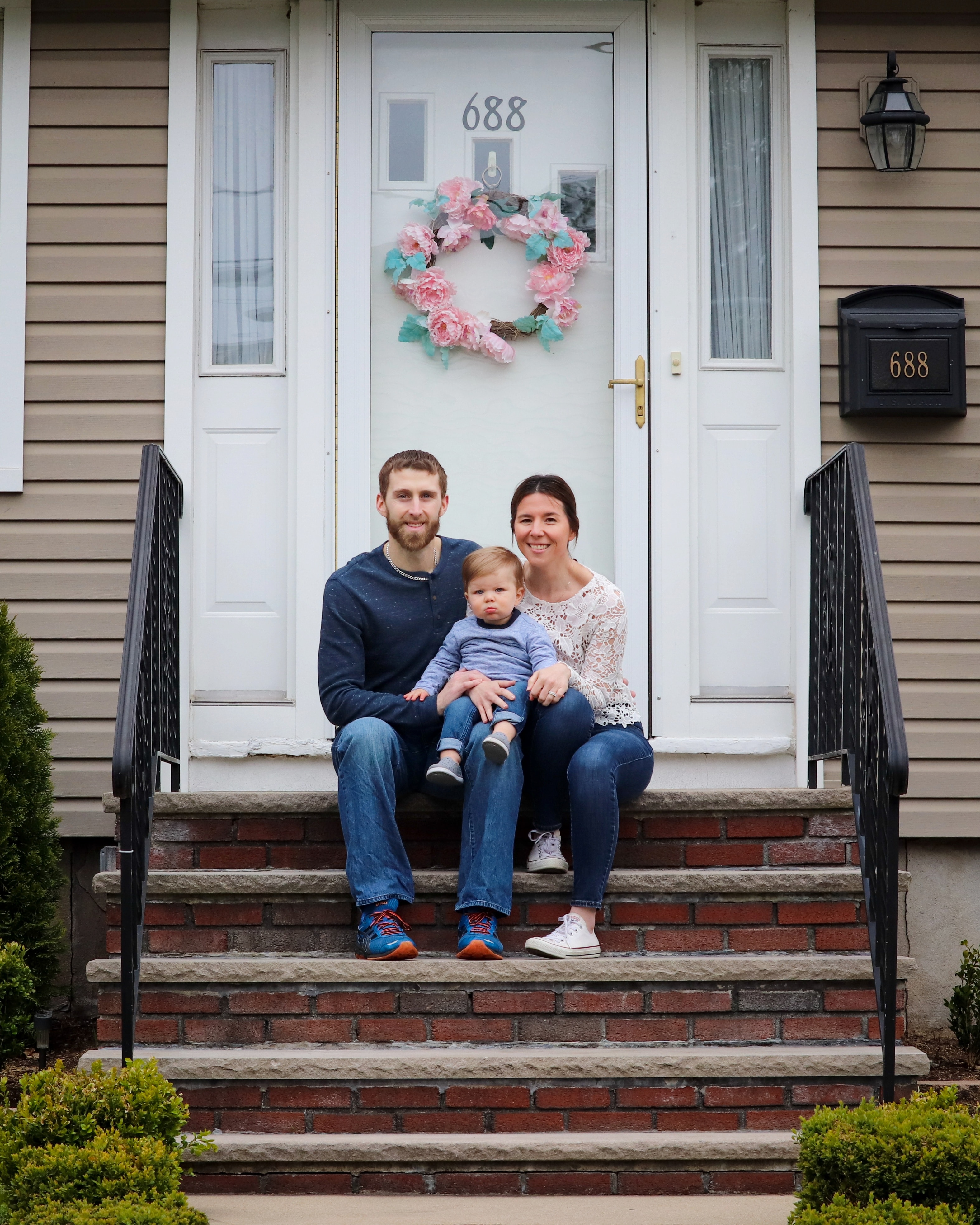 Family of three sitting in front of their front door which is white and has a wreath.