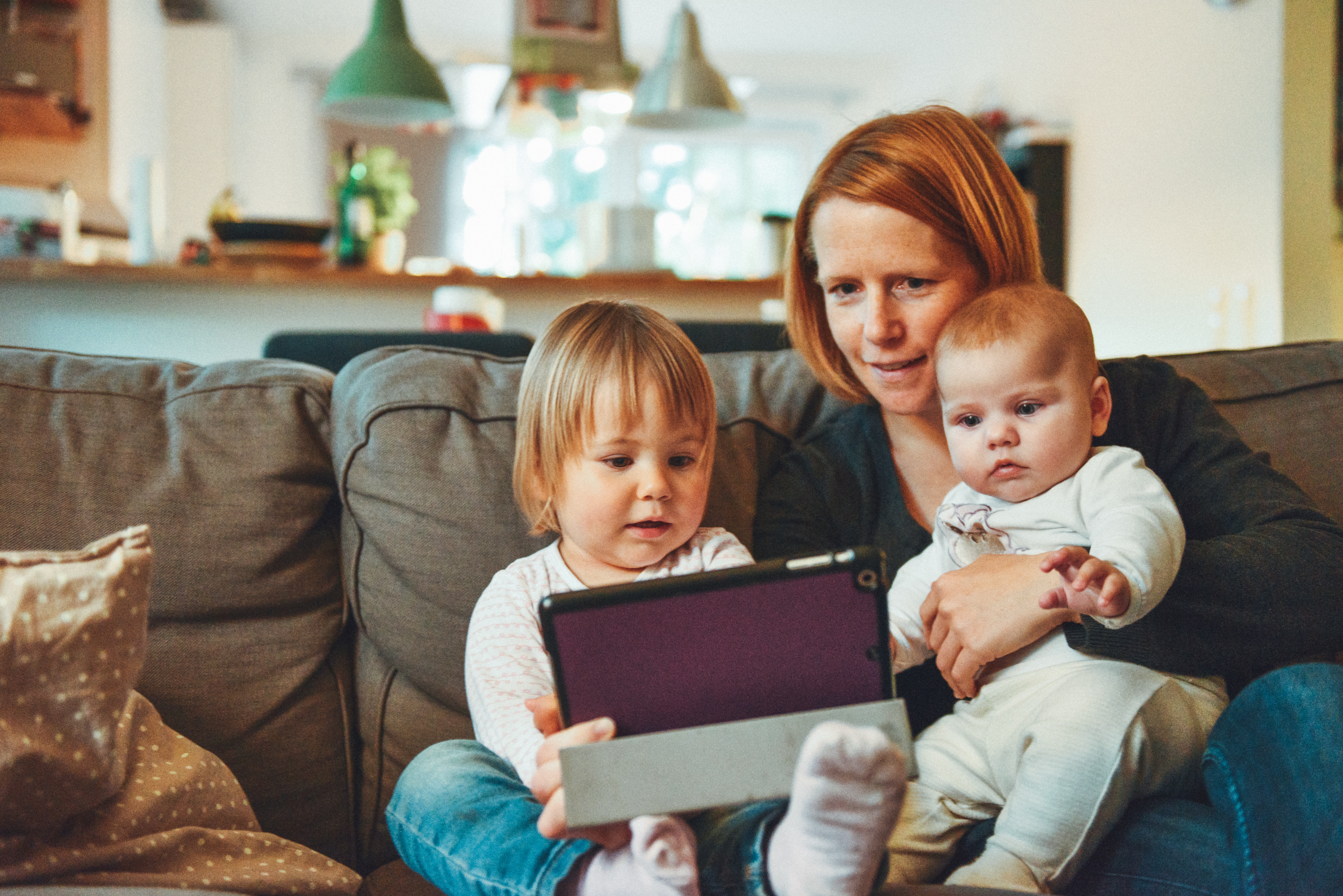 Mom with baby and toddler sitting on a couch inside their home looking at a touch screen device.